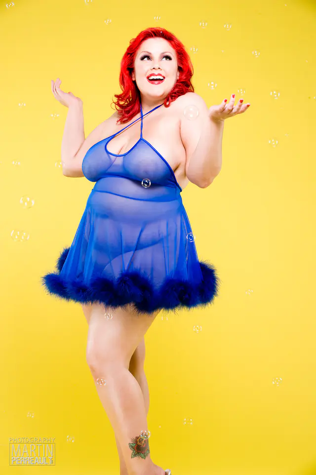A pin-up model in a blue babydoll against a yellow wall, blowing bubbles