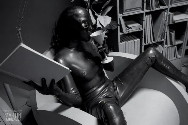 A figure in black latex reading in a library, in black and white