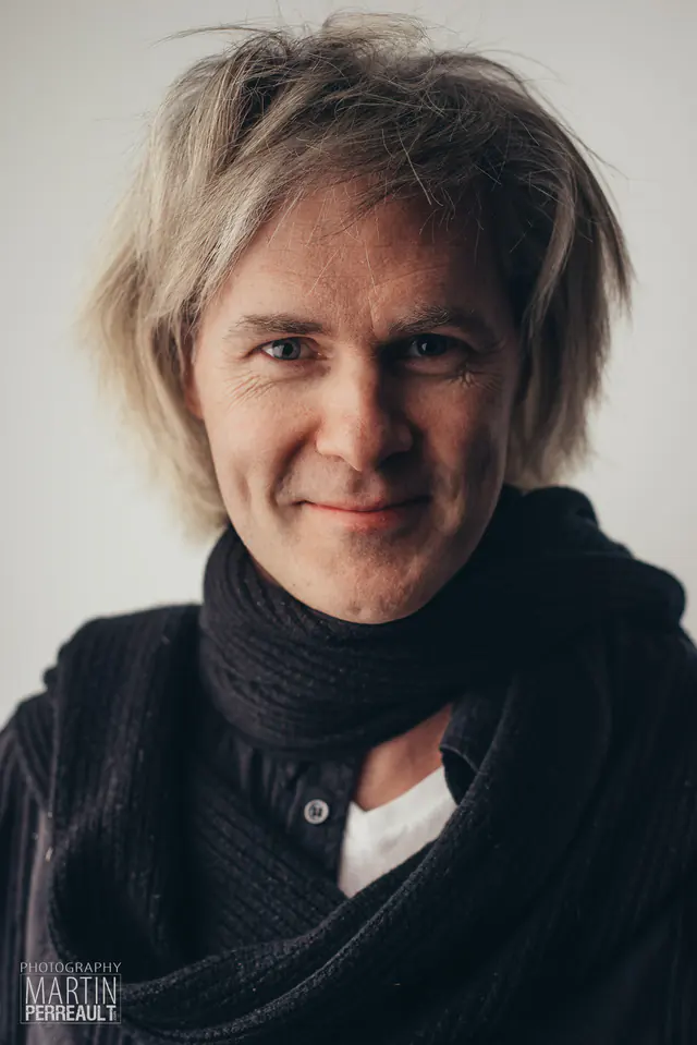 A smiling man with tousled blond hair and a black scarf
